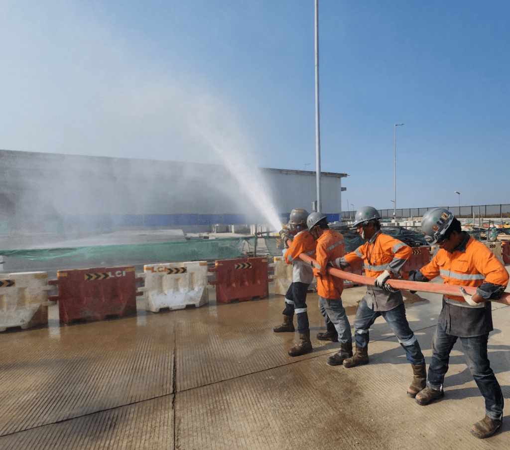 Four workers wearing orange safety uniforms, helmets, and gloves are participating in a hydrant testing, holding a high-pressure fire hose spraying water. They are standing on a concrete surface near red and white barricades, with an industrial facility and construction materials visible in the background under a clear blue sky