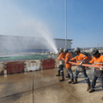 Four workers wearing orange safety uniforms, helmets, and gloves are participating in a hydrant testing, holding a high-pressure fire hose spraying water. They are standing on a concrete surface near red and white barricades, with an industrial facility and construction materials visible in the background under a clear blue sky